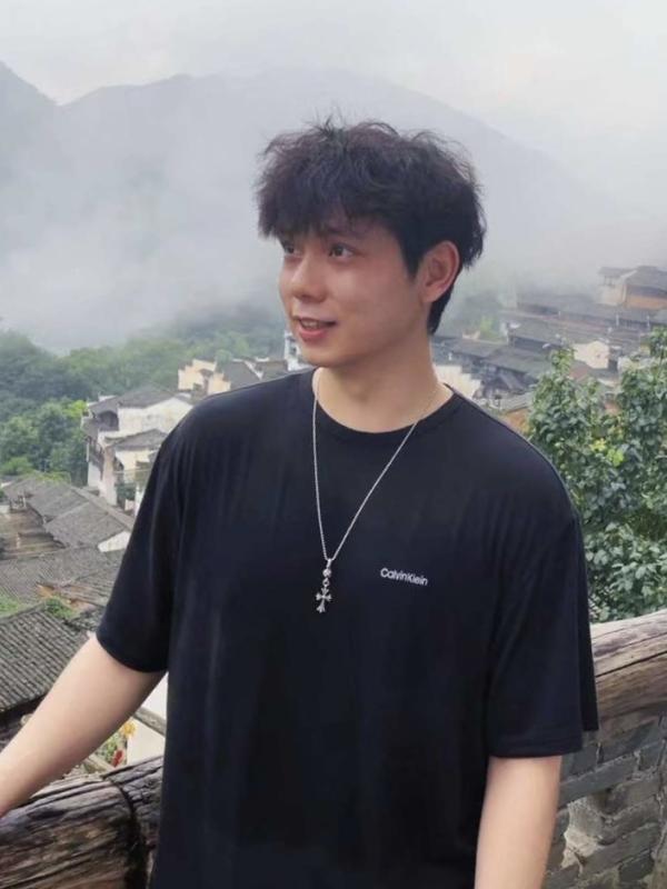 This photo features Cruz Guan smiling outdoors on a misty day. He is wearing a black Calvin Klein t-shirt and a silver necklace with a pendant. The background shows a scenic mountainous landscape with traditional buildings partially obscured by fog.