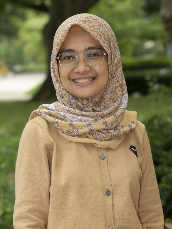 This photo features Eva Mushoffa smiling outdoors. She is wearing glasses, a beige cardigan, and a floral-patterned hijab in shades of beige and purple. The background is a lush, green park area, creating a peaceful and inviting atmosphere.