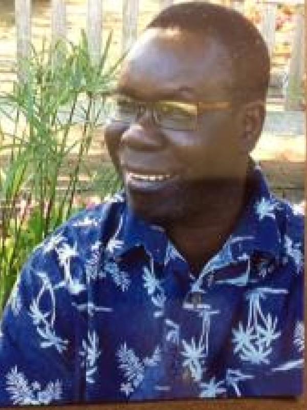 This photo features Scopas Poggo smiling outdoors. He is wearing glasses and a blue patterned shirt with white designs. The background shows a bright garden area with flowers and greenery, creating a relaxed and cheerful atmosphere.