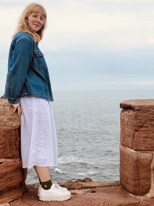 This photo features Victoria Paige smiling while standing near the edge of a rocky coastline. She is wearing a denim jacket, a white skirt, and white sneakers. The background shows the ocean and a cloudy sky, creating a serene and scenic atmosphere.
