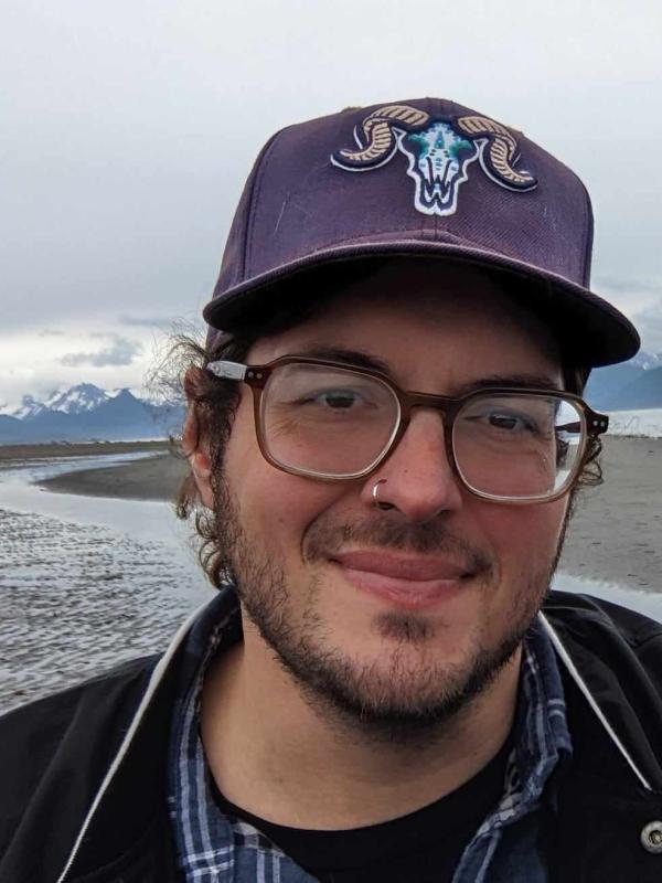 This photo features Nick Mauro smiling outdoors. He is wearing glasses, a navy blue baseball cap with a ram skull emblem, and a plaid shirt layered with a black jacket. The background is a serene coastal landscape with mountains in the distance under a cloudy sky.